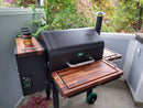 A BBQ Boards® GMG Daniel Boone/Ledge Trio Front, Side & Pellet Bin Cutting Boards (Set of Three) is a perfect BBQ prep station on the patio, with three seasoning bottles on the left shelf and trees and plants behind it.