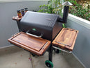 A BBQ Boards® GMG Daniel Boone/Ledge Trio Cutting Board set of three is displayed on a pellet grill with wooden side shelves and grilling accessories, serving as a BBQ prep station on a patio near a light blue wall and greenery.