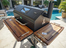 A modern outdoor grill with BBQ Boards® GMG Jim Bowie/Peak Pair—front & side cutting boards—stands on a stone patio by a pool, amid palm plants and seating in a sunny backyard.