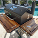 A GMG Jim Bowie Peak outdoor smoker grill, featuring BBQ Boards® Front & Side Cutting Boards (Pair), sits on a patio by a pool with a modern house and palm trees in the background. The grill has a glass window on the lid.