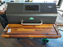 A close-up of a BBQ Boards® GMG Jim Bowie/Peak Front Cutting Board on a black pellet grill with a metal handle, featuring an accessory window. Bottles of BBQ rub and seasonings rest on the BBQ prep station side shelf.