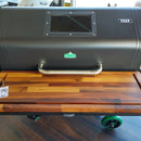 A BBQ Boards® GMG Jim Bowie/Peak Front Cutting Board by BBQ Boards® serves as a prep station on a black GMG Peak pellet grill with a glass window lid, metal handle, green wheels, and a temperature tag, all set on dark wood flooring.