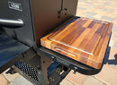 The BBQ Boards® GMG Ledge Prime 2.0 Pair, Front & Side Cutting Boards by BBQ Boards® sit on a black metal grill shelf, making an ideal upgrade for your Green Mountain Grills setup, with patio pavers seen in the background.