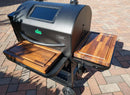 A black outdoor pellet grill with two wooden side shelves, each fitted with a BBQ Boards® GMG Ledge Prime 2.0 Pair (front & side cutting boards), sits on a brick patio in the sunlight.