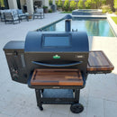 A large black pellet grill with wooden side shelves features BBQ Boards® GMG Ledge Prime 2.0 Pair Front & Side Cutting Boards, sitting on a stone patio by a pool, surrounded by outdoor seating and lush greenery.