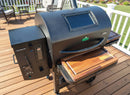 A black pellet grill with a digital panel features BBQ Boards® GMG Ledge Prime 2.0 Front & Side Cutting Boards (Pair) on its shelves, set on a wooden deck with patio furniture and railing—ideal for your outdoor Green Mountain Grills BBQ setup.