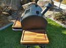 A black outdoor smoker grill with a chimney and two wooden side shelves stands on grass, featuring the BBQ Boards® GMG Ledge Prime 2.0 Pair—Front & Side Cutting Boards by BBQ Boards®. Rocks and plants provide a scenic backdrop.