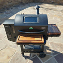 A black outdoor pellet grill on wheels with wooden shelves features the BBQ Boards® GMG Ledge Prime 2.0 Pair, Front & Side Cutting Boards—the perfect Green Mountain Grills accessory to upgrade your setup, shown on a stone patio against a stone wall.