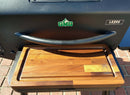 A BBQ Boards® GMG Ledge Prime 2.0 Front Cutting Board sits on the front shelf of a black pellet grill displaying the Green Mountain Grills LEDGE logo, with both products resting on a brick surface.