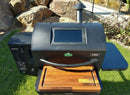 A black GMG Ledge Prime 2.0 pellet grill, featuring the BBQ Boards® GMG Ledge Prime 2.0 Front Cutting Board by BBQ Boards®, sits on grass with rocks and plants in the background, complemented by Green Mountain Grills accessories.