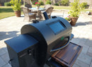 A modern outdoor pellet grill with a digital display is on a stone patio, featuring the BBQ Boards® GMG Ledge Prime 2.0 Front Cutting Board, with potted plants and a patio dining set in the background.