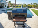 A black outdoor pellet grill and smoker with a BBQ Boards® GMG Ledge Prime 2.0 Front Cutting Board is set on a stone patio beside a modern pool, with palm trees and contemporary houses in the background.