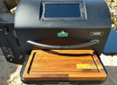 A black pellet grill with a digital control panel features the BBQ Boards® GMG Ledge Prime 2.0 Front Cutting Board by BBQ Boards®. This Green Mountain Grills setup is outdoors on a sunny day.
