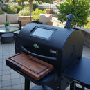 A black pellet grill with a BBQ Boards® GMG Ledge Prime 2.0 Front Cutting Board stands on a stone patio, surrounded by outdoor furniture and greenery in a sunny backyard.