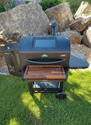 A black pellet grill is on green grass, featuring the BBQ Boards® GMG Ledge Prime 2.0 Front Cutting Board by BBQ Boards® on its side shelf, with rocks and bushes in the background—a perfect outdoor cooking setup with must-have accessories.