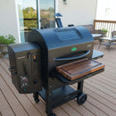 A BBQ Boards® GMG Ledge Prime 2.0 Front Cutting Board is installed on a black outdoor pellet grill with a digital control panel, set on a wooden deck amid patio furniture and potted plants. Green Mountain Grills accessories complete the look.