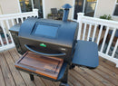 A black pellet grill with a windowed lid and digital controls sits on a deck. A BBQ Boards® GMG Ledge Prime 2.0 Front Cutting Board is on the front board, with Green Mountain Grills accessories and patio furniture in the background.