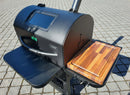 A black outdoor smoker grill with a metal shelf on the left and a BBQ Boards® GMG Ledge Prime 2.0 Side Cutting Board as a BBQ prep station on the right, standing on a paved surface.
