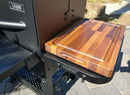 The BBQ Boards® GMG Ledge Prime 2.0 Side Cutting Board sits on a black grill’s side shelf, creating an ideal BBQ prep station, with sunlight casting shadows across the paved patio.