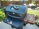 A BBQ Boards® GMG Ledge Prime 2.0 Side Cutting Board sits on a backyard patio, creating a perfect BBQ prep station amid flowers and outdoor furniture on a sunny day.