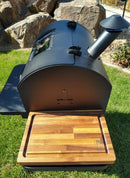 A black outdoor smoker grill with a side chimney stands on grass. In front, the BBQ Boards® GMG Ledge Prime 2.0 Side Cutting Board by BBQ Boards® with grooves and logo is displayed, with rocks and plants in the background.