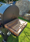 A close-up of a BBQ Boards® GMG Ledge Prime 2.0 Side Cutting Board attached to a black outdoor grill, set on green grass with rocks and plants—a perfect Green Mountain Grills accessory for your BBQ prep station.