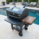 A black pellet grill with a BBQ Boards® GMG Ledge Prime 2.0 Side Cutting Board doubles as a prep station on a poolside patio, surrounded by outdoor furniture and potted plants.