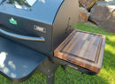 A black pellet grill features the BBQ Boards® GMG Ledge Prime 2.0 Side Cutting Board, creating an ideal BBQ prep station on green grass with large rocks in the background.