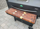 A pair of BBQ Boards® GMG Peak Prime 2.0 Front & Side Cutting Boards by BBQ Boards® is mounted on a black GMG PEAK grill, creating a convenient workspace on the stone patio.