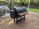 A BBQ Boards® GMG Peak Prime 2.0 Pair, Front & Side Cutting Boards (Pair) is attached to a black outdoor pellet grill on a wooden deck, surrounded by patio furniture, plants, green lawn, and trees for a relaxing setting.