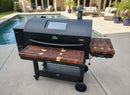 The BBQ Boards® GMG Peak Prime 2.0 Pair, Front & Side Cutting Boards by BBQ Boards® provide extra workspace for your black outdoor pellet grill on the patio, with couches and potted plants visible near a swimming pool.