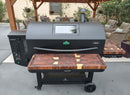 A BBQ Boards® GMG Peak Prime 2.0 cutting board pair sits on a large outdoor pellet grill with a glass lid window beneath a wooden pergola, surrounded by side shelves, chairs, plants, and a butcher block table on the patio.