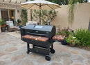 A spacious stone patio, with potted plants and outdoor furniture under a beige umbrella, features a large black pellet grill equipped with BBQ Boards® GMG Peak Prime 2.0 Front & Side Cutting Boards (Pair) for added convenience.