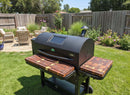 On a sunny day, BBQ Boards® GMG Peak Prime 2.0 Pair Front & Side Cutting Boards by BBQ Boards® add extra prep space to your backyard grill setup, surrounded by patio furniture, a wooden fence, and flowering plants on green grass.