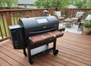 On a wooden deck near outdoor furniture, the sleek GMG Peak Prime 2.0 pellet grill is paired with BBQ Boards® Front & Side Cutting Boards (Pair), offering modern style, digital controls, and spacious prep space—perfect for Green Mountain Grills accessories.