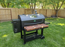 A large black pellet grill fitted with a BBQ Boards® GMG Peak Prime 2.0 Front Cutting Board stands on backyard grass, surrounded by a wooden fence, trees, and two black patio chairs in the background.