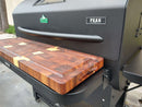 A close-up of a GMG Peak grill featuring the BBQ Boards® GMG Peak Prime 2.0 Front Cutting Board on its shelf, providing a premium prep space for grilling outdoors on a concrete surface.