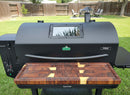 A large black pellet grill features a BBQ Boards® GMG Peak Prime 2.0 Front Cutting Board, providing ample prep space. It sits on a green lawn with backyard views, fencing, and patio furniture visible in the background.