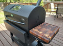 A large black outdoor pellet grill with a BBQ Boards® GMG Peak Prime 2.0 Side Cutting Board sits on a wooden deck, surrounded by patio furniture, Green Mountain Grills accessories, and green backyard trees.