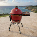 A BBQ Boards® Kamado Joe Classic Joe I Side Cutting Board is set on a tiled patio, overlooking the scenic coastline with cliffs and ocean waves, offering outdoor cooks extra prep space by their grill.