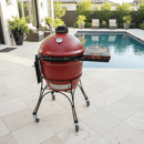 A BBQ Boards® Kamado Joe Classic Joe I Side Cutting Board from BBQ Boards® is attached to a red ceramic grill on wheels, providing prep space by the pool among green plants and outdoor seating.