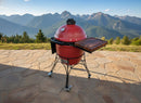 A BBQ Boards® Kamado Joe Classic Joe I Side Cutting Board is set up on a stone patio with a red ceramic grill, overlooking scenic mountains and a green forest under a clear blue sky.