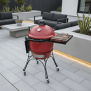 A red ceramic kamado grill with a BBQ Boards® Kamado Joe Classic Joe I Side Cutting Board on wheels stands on a modern patio with grey tiles, outdoor seating, and potted plants, providing convenient BBQ prep space.
