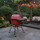 A BBQ Boards® Kamado Joe Classic Joe I Side Cutting Board is paired with a red ceramic grill on a patio under a wooden pergola with string lights, surrounded by cozy outdoor seating, lush greenery, and inviting lighting.