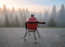 A red ceramic grill with a BBQ Boards® Kamado Joe Classic Joe I Side Cutting Board stands on a stone patio overlooking a misty evergreen forest at sunrise or sunset.