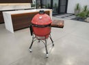A red ceramic kamado grill with a thermometer and BBQ Boards® Kamado Joe Classic Joe I Side Cutting Board by BBQ Boards®, plus wheels, is on a modern patio near a white countertop and green plants.