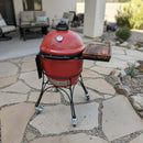 A red Kamado-style BBQ grill with built-in thermometer sits on a stone patio. It features a BBQ Boards® Kamado Joe Classic Joe I Side Cutting Board as prep space, black metal legs, wheels, and is surrounded by outdoor furniture and plants.