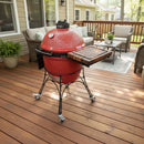 A BBQ Boards® Kamado Joe Classic Joe I Side Cutting Board by BBQ Boards® sits atop a red ceramic outdoor grill on a wooden deck, surrounded by patio furniture and potted plants, creating the perfect BBQ prep space.