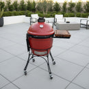 A red ceramic Kamado-style grill with a BBQ Boards® Kamado Joe Classic Joe I Side Cutting Board stands on a gray tiled patio, surrounded by modern outdoor furniture and green shrubs in the background.