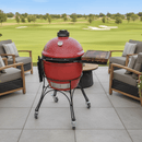 A red Kamado Joe Classic Joe I sits on a patio with wicker chairs and cushions. Next to it, a BBQ Boards® Kamado Joe Classic Joe I Side Cutting Board provides BBQ grill prep space. A golf course lies beyond under the blue sky and trees.